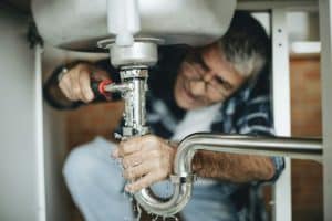 expert Residential Plumbing and Emergency Services in Northeast Indiana | adult repairman with red plastic bucket holding pipes for repairing kitchen sink Plumber fixing the kitchen sink